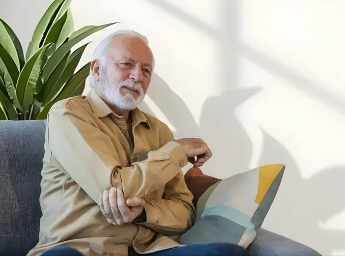 Older man on a sunlit sofa cradling his sore forearm and elbow